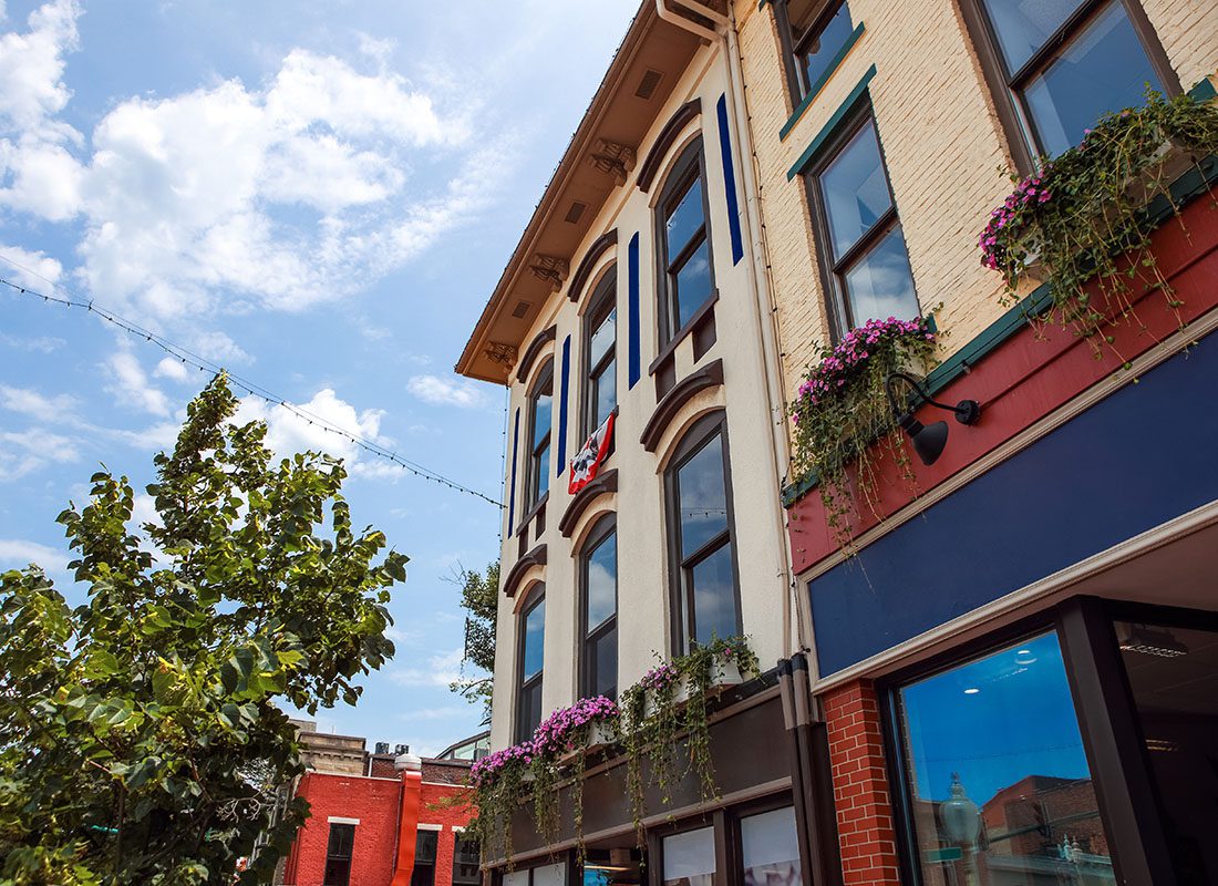 Contact - Angled View Looking Up at Historical Brick Buildings with Flowers on the Windows and Small Business Shops Downstairs Against a Cloudy Blue Sky in a Small Indiana Town